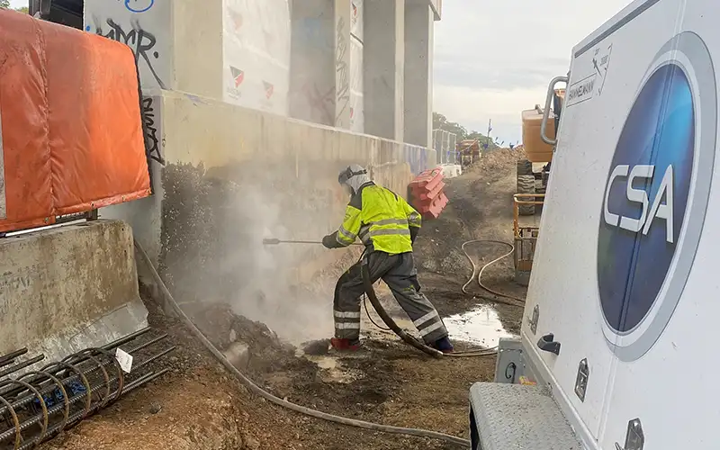 Worker in high-visibility clothing using a water jetting lance beside CSA service equipment at an industrial site